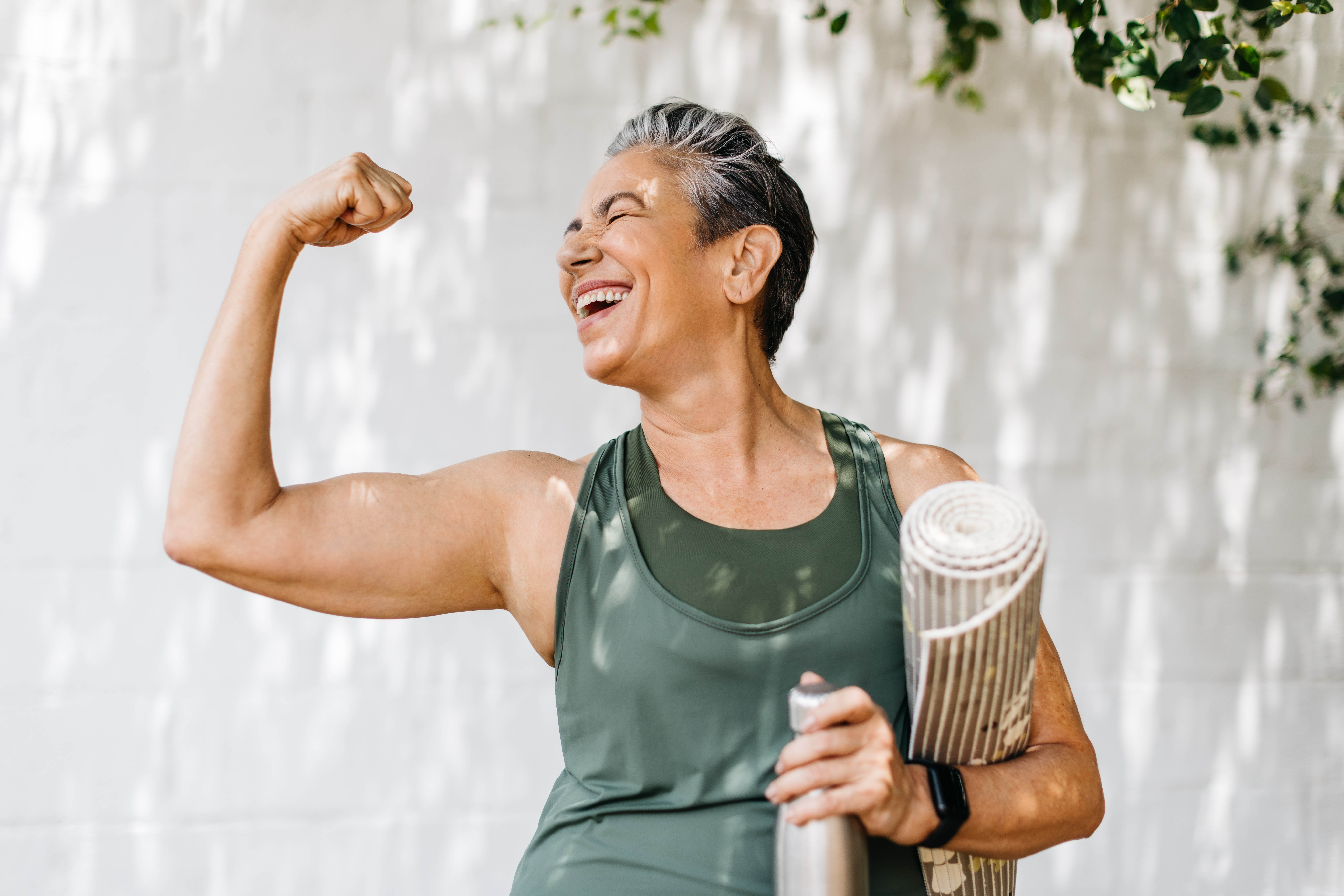 Older woman flexing her arm and smiling, she has a yoga mat under her other arm